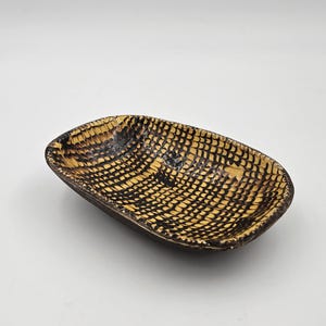 May include: An oval-shaped ceramic dish featuring a textured interior with a repeating black grid pattern over a yellow base. The exterior of the bowl is dark brown, and the rim is slightly uneven, showcasing a handcrafted appearance and finish.