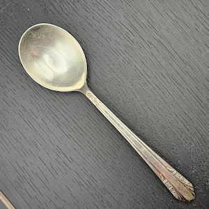 May include: A silver spoon with an oval bowl and a long, decorated handle. The spoon is made of a shiny, reflective metal and rests on a dark, textured surface. The handle features a decorative pattern near the bowl and at the end.