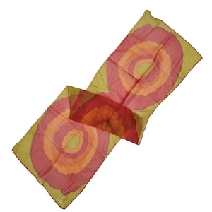 May include: A rectangular scarf with an abstract design. It features concentric circles in red, orange, and yellow on a pale green background. The word "Vera" is printed in the corner of the scarf.