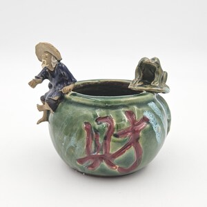 Peut inclure: Un pot en céramique émaillée verte de forme ronde, orné d'une figurine sculptée représentant une personne portant un chapeau beige et des vêtements bleus assise sur le bord, ainsi qu'une petite grenouille verte. Le devant présente des caractères calligraphiés rouge foncé.