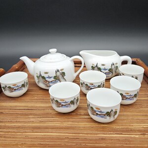 Könnte beinhalten: Ein weißes Porzellan-Tee-Set mit Landschaftsdesign. Das Set enthält eine Teekanne, einen Milchkännchen und sechs Teetassen. Die Teetassen und die Teekanne haben ein blaues und grünes Landschaftsdesign auf weißem Hintergrund. Der Milchkännchen hat ein blaues und grünes Landschaftsdesign auf weißem Hintergrund. Das Set befindet sich auf einer Bambusmatte.