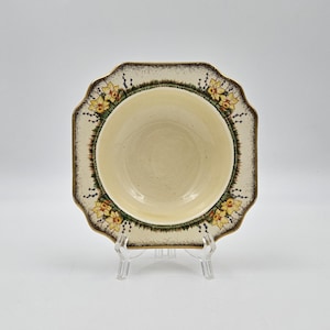 May include: A vintage ceramic bowl with a square shape and scalloped edges. The bowl has a cream-coloured interior and a decorative border with yellow floral patterns and a brown trim. The bowl is displayed on a clear plastic stand.