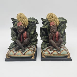 May include: A pair of decorative bookends featuring a monkey figure wearing a red vest and hat, seated on a patterned pillow. The monkey is set against a backdrop of green leaves and a cluster of bananas. The base is black.