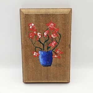 Könnte beinhalten: Rechteckige Holzplakette mit handgemaltem Blumendesign. Das Kunstwerk zeigt rote Blumen und grüne Stiele in einer blauen Vase. Der Hintergrund ist eine natürliche Holzmaserung.
