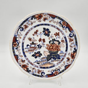May include: A white ceramic plate with a scalloped edge features a blue, red, and gold floral design. The center of the plate depicts a pagoda-like structure with a floral motif.