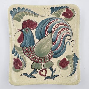 May include: A square ceramic plate with a rooster design. The rooster is painted in shades of blue, green, and red, with a cream-colored background. The plate has a slightly cracked appearance, adding to its vintage charm.