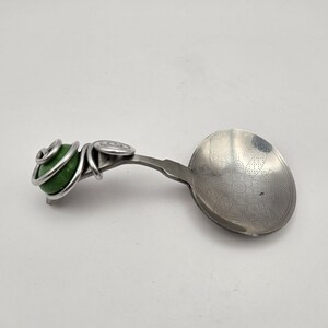 May include: A decorative spoon with a green stone handle wrapped in silver-colored wire. The spoon's bowl has an etched floral design. The spoon is set against a white background.