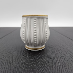 May include: A small, white ceramic vase with a textured, ribbed design and a gold rim. The vase features vertical grooves and a series of small, raised dots. The base also has a gold accent. The vase is a decorative item.