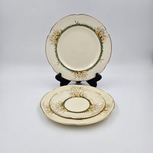 May include: A set of three cream-colored ceramic plates with a floral design. The plates have a scalloped edge with a gold trim. The floral design features yellow flowers and green leaves. The plates are displayed on a black plate stand.