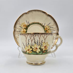 Könnte beinhalten: Cremefarbenes Tee-Set mit floralem Design. Die Tasse und Untertasse zeigen gelbe Narzissen, grünes Gras und braune Äste. Die Ränder sind goldfarben verziert. Die Untertasse hat einen gewellten Rand.