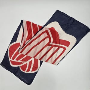 May include: A rectangular scarf featuring a dark navy blue background with a large, stylized red and cream-colored graphic print. The design consists of bold, curved red lines on a cream base, and the name Adele Simpson is printed in the corner.