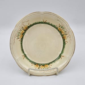 May include: Cream-colored ceramic plate with a scalloped edge and a gold rim. The plate features a green border with yellow daffodil floral design. The plate is displayed on a white metal stand.