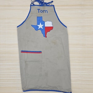 May include: A light grey apron featuring blue trim and ties. The name Tom is embroidered in blue at the top. Below the name is a graphic of the state of Texas in blue, white, and red. A single rectangular pocket with a blue and red trim is present.