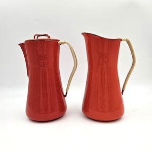 May include: A set of two red metal pitchers with a glossy finish. One pitcher features a lid with a small handle, while both vessels are equipped with curved handles wrapped in light-colored, textured material for a distinct grip and appearance.