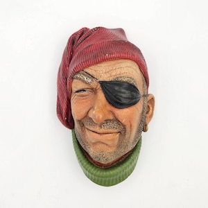 May include: A wall-mounted decorative sculpture of a face wearing a red knit cap and a green ribbed collar. The figure has a black eye patch over the left eye, a light brown complexion with visible skin texture, and a small gold hoop earring.