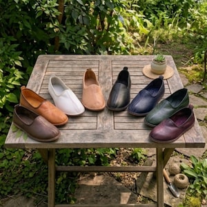 Puede incluir: Una colección de zapatos sin cordones de cuero en varios colores, incluyendo marrón, blanco, negro y azul, dispuestos sobre una mesa de madera. Los zapatos se exhiben al aire libre, rodeados de vegetación, sugiriendo un entorno natural.