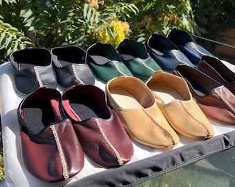 May include: A collection of nine leather slippers in various colors, including black, green, brown, and beige. The slippers are arranged in a row on a black surface.