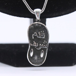 May include: Silver pendant necklace with a detailed design of a footprint with Arabic calligraphy. The inscription reads "Wadam Bashir".