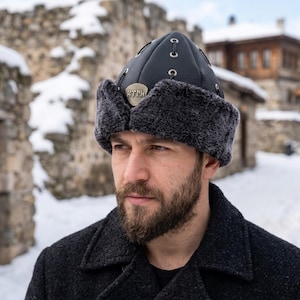 Può includere: Un cappello in pelle nera con finiture in pelliccia grigia e un'emblema metallico. Il cappello ha una parte superiore arrotondata con cuciture decorative e viene indossato in un ambiente esterno innevato. Parte di una collezione di abbigliamento invernale.