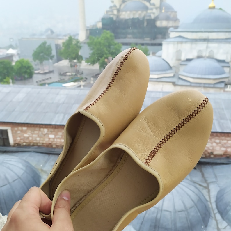 May include: A pair of beige leather slippers with brown stitching. The slippers are worn and have a slightly distressed look.