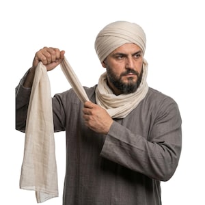 Puede incluir: Un hombre con un turbante color crema y una bufanda a juego. Sostiene el extremo de la bufanda, que está drapeada alrededor de su cuello. Lleva una camisa gris de manga larga. El fondo es blanco liso.