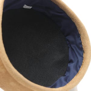 May include: Close-up of a tan suede hat with a black interior and a navy blue satin lining. The hat's interior is a dark, textured fabric. The hat is a classic style, suitable for various occasions.