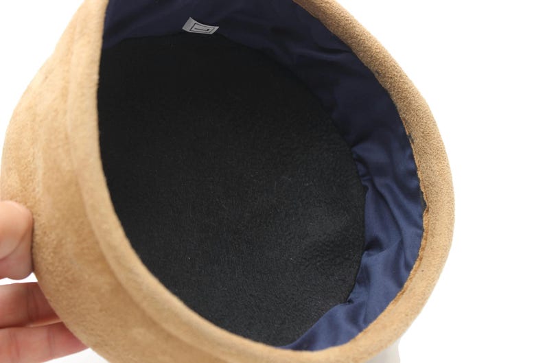 May include: Close-up of a tan suede hat with a black interior and a navy blue satin lining. The hat's interior is a dark, textured fabric. The hat is a classic style, suitable for various occasions.