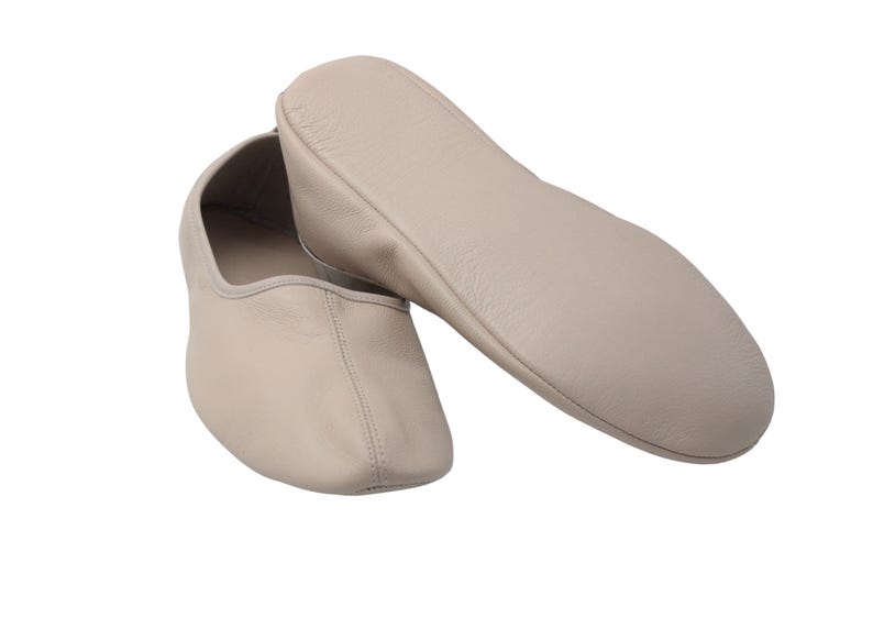 Pu&ograve; includere: Un paio di scarpette da ballo in pelle beige, una in piedi e l'altra sdraiata su un lato. Le scarpette hanno una punta arrotondata e una cucitura cucita al centro. Le suole sono lisce e la pelle sembra morbida.