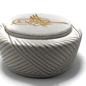May include: A white and gold turban-style hat with a rounded top and a striped, angled design on the sides. The top features a gold embroidered emblem. The hat is likely a cultural or religious head covering.