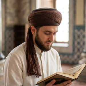 May include: A man wearing a brown turban and a white shirt is reading a book with gold-coloured pages. The man has a beard and is looking down at the book. The background is a blurred interior with pillars and patterned walls.