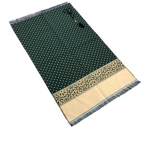 May include: A dark green prayer rug with a repeating pattern of small, light-colored dots. The rug has a decorative border with geometric designs in gold and tan, and a fringed edge. A set of prayer beads rests on the rug.