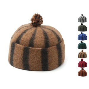 Puede incluir: Un gorro de punto marrón y negro a rayas con un pompón marrón en la parte superior. El gorro está disponible en una variedad de colores.