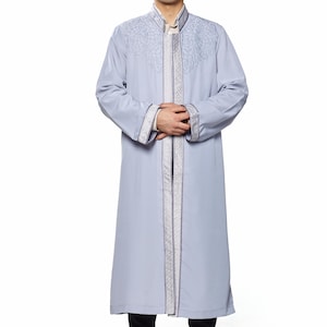 May include: A man wearing a grey robe with silver embroidery and a white hat with a red trim. He is standing with his hands clasped in front of him.