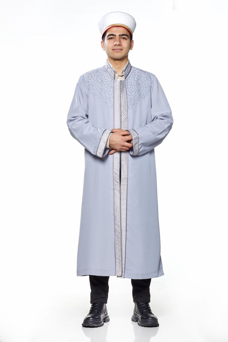 May include: A man wearing a grey robe with silver embroidery and a white hat with a red trim. He is standing with his hands clasped in front of him.