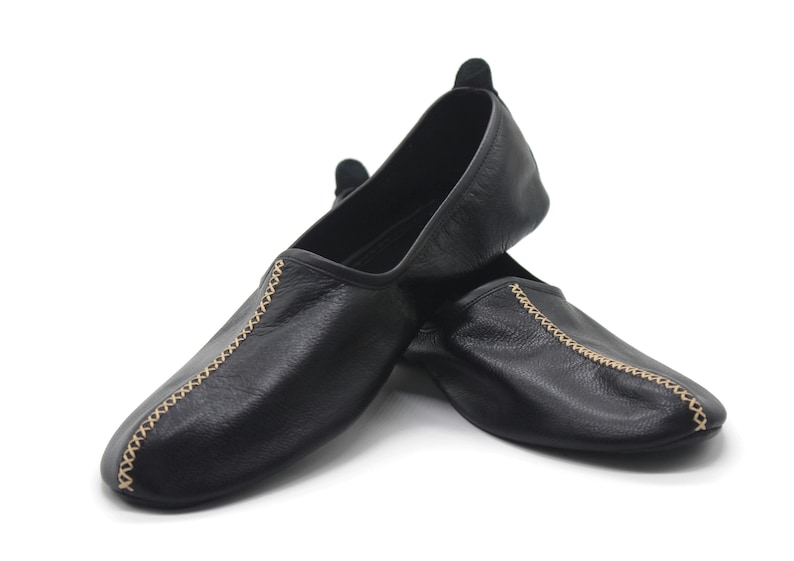 Puede incluir: Un par de zapatillas de cuero negro con costuras doradas. Las zapatillas est&aacute;n dobladas una sobre la otra.