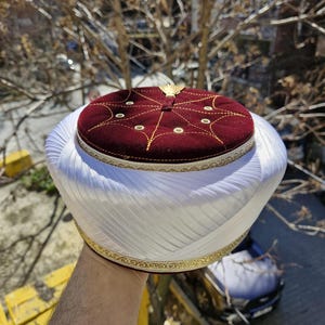 May include: A white and gold turban style hat with a maroon velvet top. The top has gold stitching and a gold button.