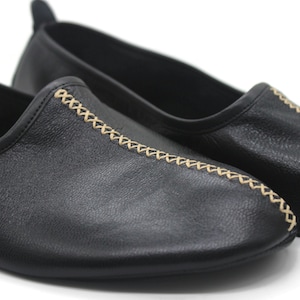 Puede incluir: Un par de zapatos negros de cuero sin cordones con un detalle de costura decorativo de color beige claro.