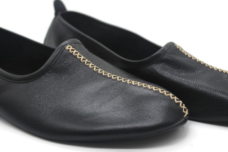 Puede incluir: Un par de zapatos negros de cuero sin cordones con un detalle de costura decorativo de color beige claro.