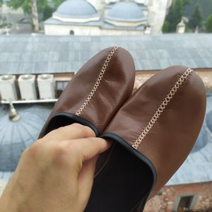 May include: A pair of brown leather slippers with a decorative stitch detail. The slippers have a black sole and are made of soft, supple leather.