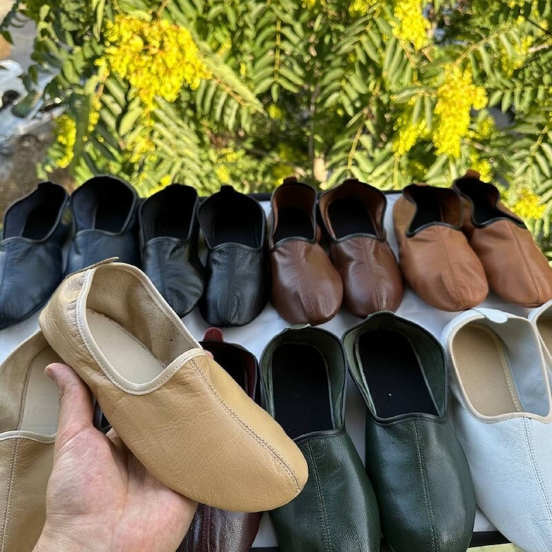 May include: A variety of leather slippers in different colors, including black, brown, beige, green, and white. The slippers are lined with a soft, white fabric.