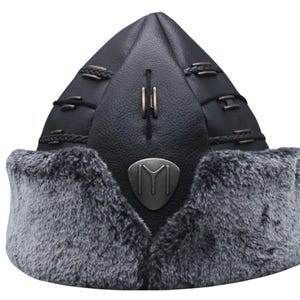 May include: A black leather hat with a grey fur trim and a metal emblem featuring the letter "M" in a stylised design.