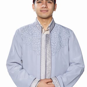 May include: A man wearing a light blue robe with intricate embroidery and a white hat with a red trim. The robe has a high collar and long sleeves.