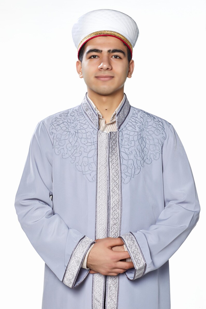 May include: A man wearing a light blue robe with intricate embroidery and a white hat with a red trim. The robe has a high collar and long sleeves.