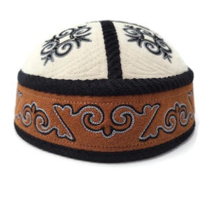 Peut inclure: Une toque ronde avec un sommet crème orné de broderies noires en volutes et une bande marron décorée de motifs tourbillonnants gris foncé et noirs. La toque est rehaussée d'un épais galon tressé noir le long des coutures et du bord inférieur.