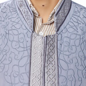May include: A grey embroidered shirt with a braided trim around the collar and down the front. The shirt has a button closure and a light brown striped undershirt is visible.