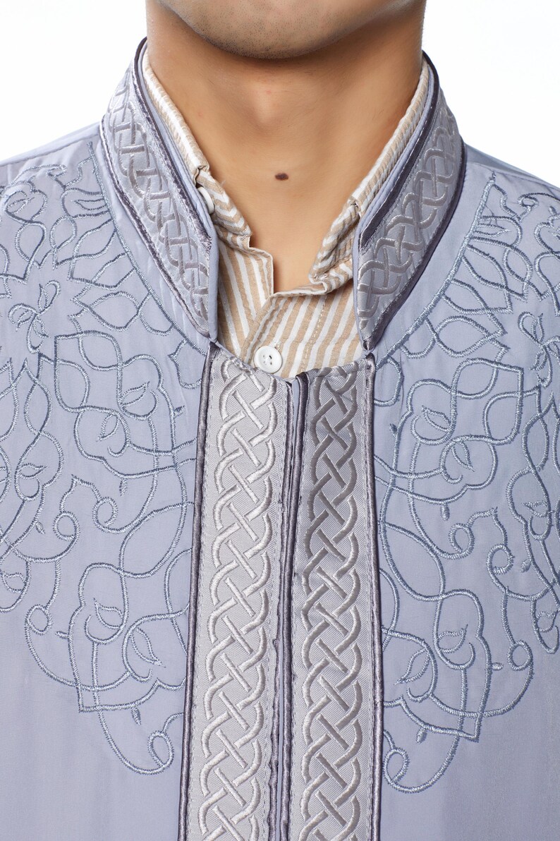 May include: A grey embroidered shirt with a braided trim around the collar and down the front. The shirt has a button closure and a light brown striped undershirt is visible.
