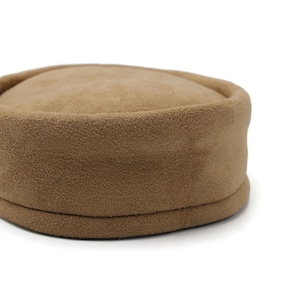 May include: A tan fleece hat with a rounded crown and a flat, circular brim. The hat is a solid color and appears to be made of a soft, textured material. The hat is a neutral color and could be worn by anyone.