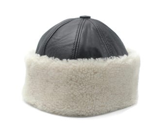 May include: A black leather hat with a rounded crown and a fluffy, off-white shearling brim. The hat has a small, round button on top and is designed to keep the wearer warm in cold weather.