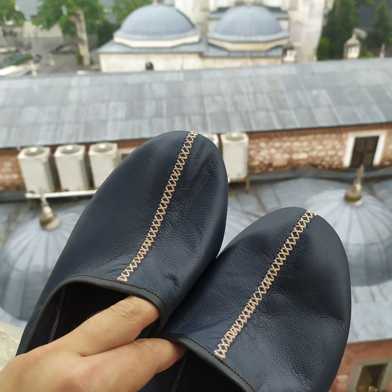 May include: A pair of dark blue leather slippers with a decorative tan stitching detail. The slippers are worn and have a slightly distressed look.
