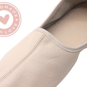 Pu&ograve; includere: Primo piano di una ballerina color crema. La scarpa &egrave; realizzata in pelle e presenta una cucitura cucita al centro. Una grafica circolare rosa con le parole "HANDMADE WITH LOVE" circonda un cuore rosa.